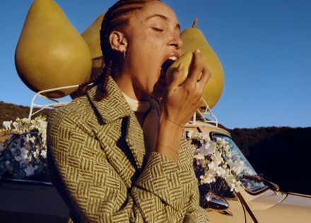 Adwoa Aboah with braided hair, biting into a pear with backdrop of three large pear props placed on top of a car roof rack.