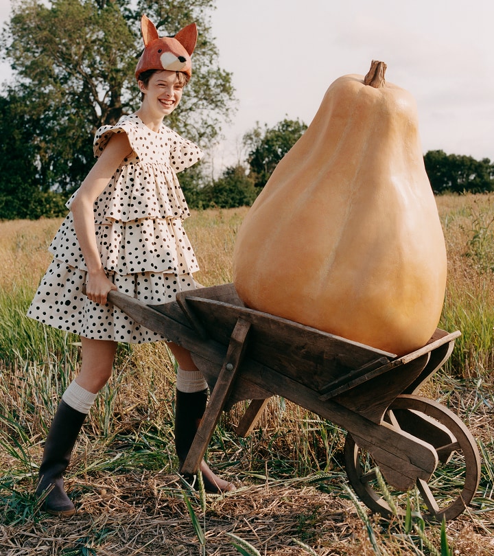 Girl in a frill dress with wheel barrow carrying a extra large butternut vegetable prop in the middle of a farm.