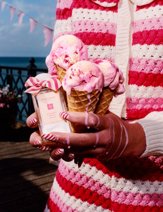 Three ice cream cones held next to a Jo Malone London cologne adorned with a pink bow, set against a scenic waterfront pier.