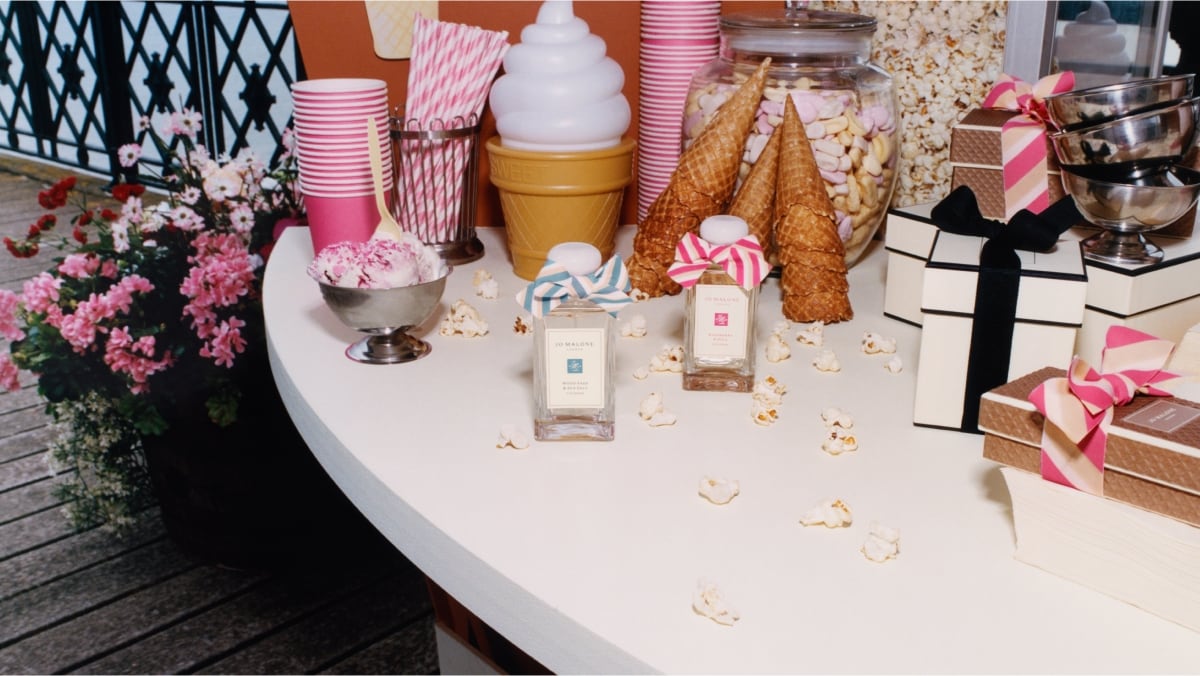 Two Jo Malone London colognes, one with a blue bow, one with a pink bow surrounded by pink cups and straws, popcorn and sweets. 