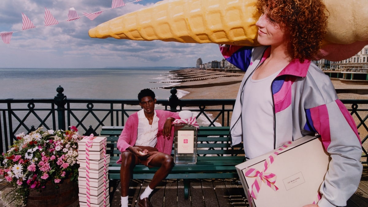 A person holding an oversized icecream walks past a person on a bench next to an oversized cologne beside a stack of gift boxes.