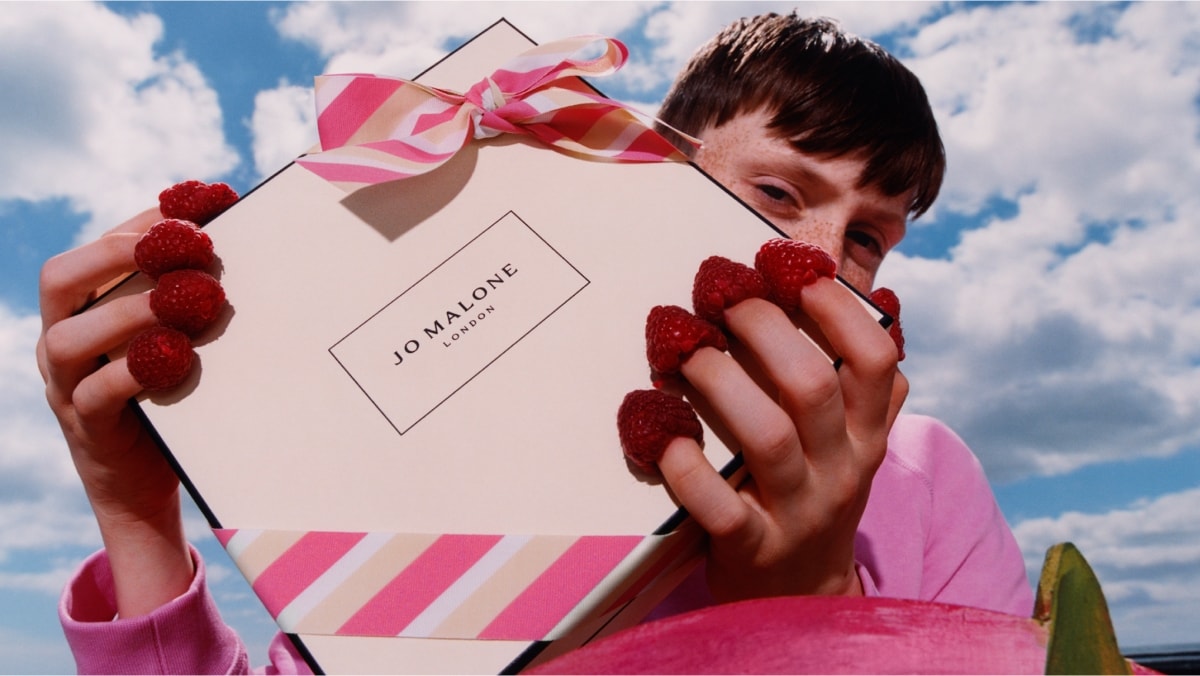 A Jo Malone London box with a pink striped bow held by a person who has raspberries on tips of their fingers, against a blue sky