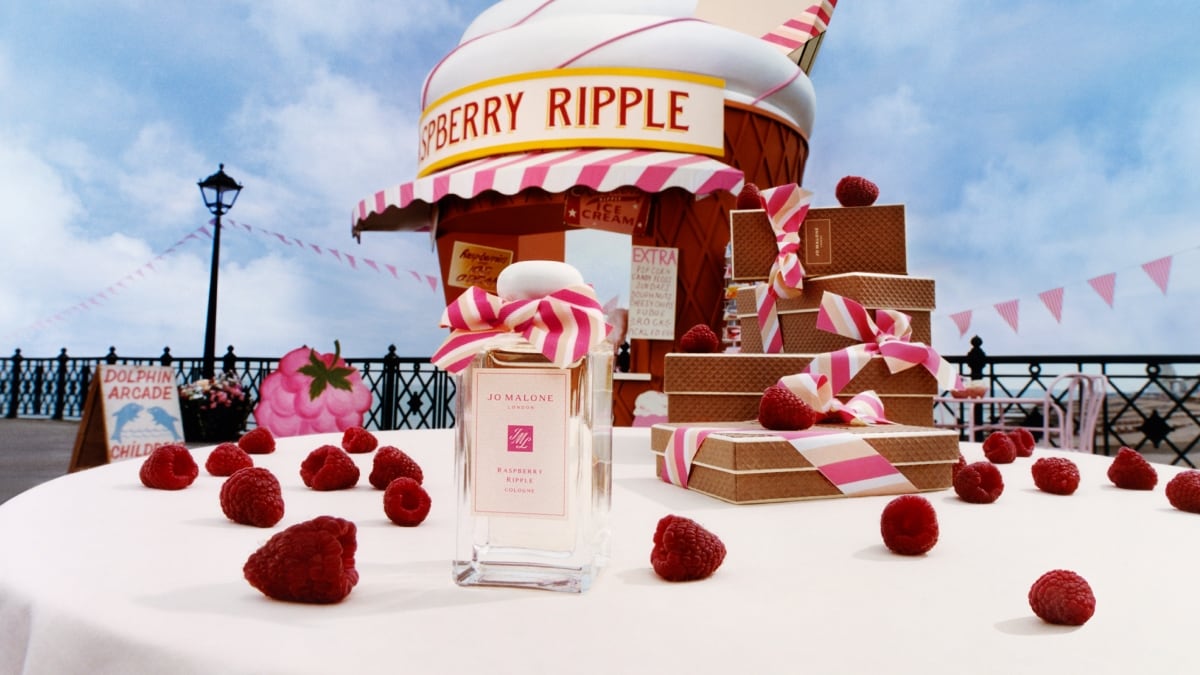 A Jo Malone London cologne with a pink bow surrounded by raspberries and a giant cone hut next to stacked waffle gift boxes.