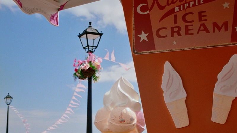 A person holds an icecream whilst walking along a pier full of people and vibrant beach props before dropping the icecream, 
