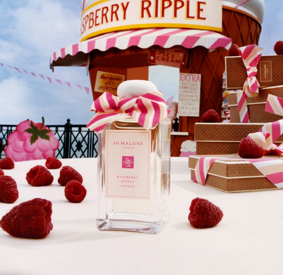 A Jo Malone London cologne with a pink bow surrounded by raspberries and a giant cone hut next to stacked waffle gift boxes.