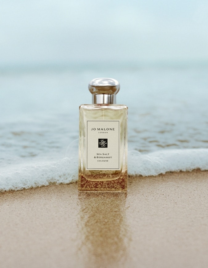 Jo Malone London Sea Salt & Bergamot cologne standing on wet sand with gentle waves behind.