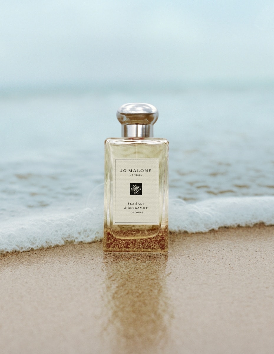 Jo Malone London Sea Salt & Bergamot cologne standing on wet sand with gentle waves behind.