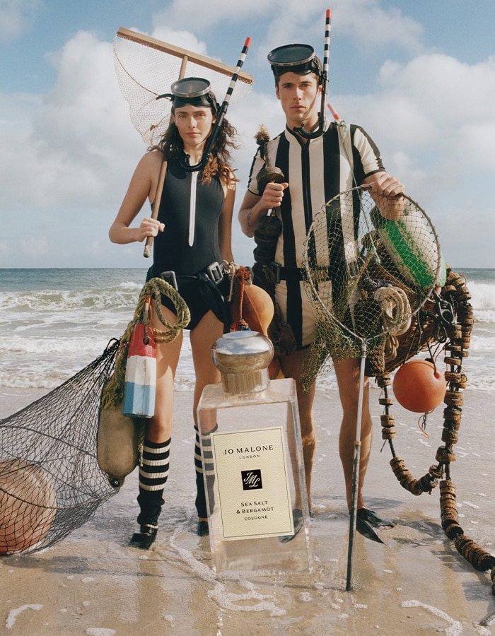 Two people in swimwear hold nets and buoys on beach with oversized Jo Malone London cologne bottle.