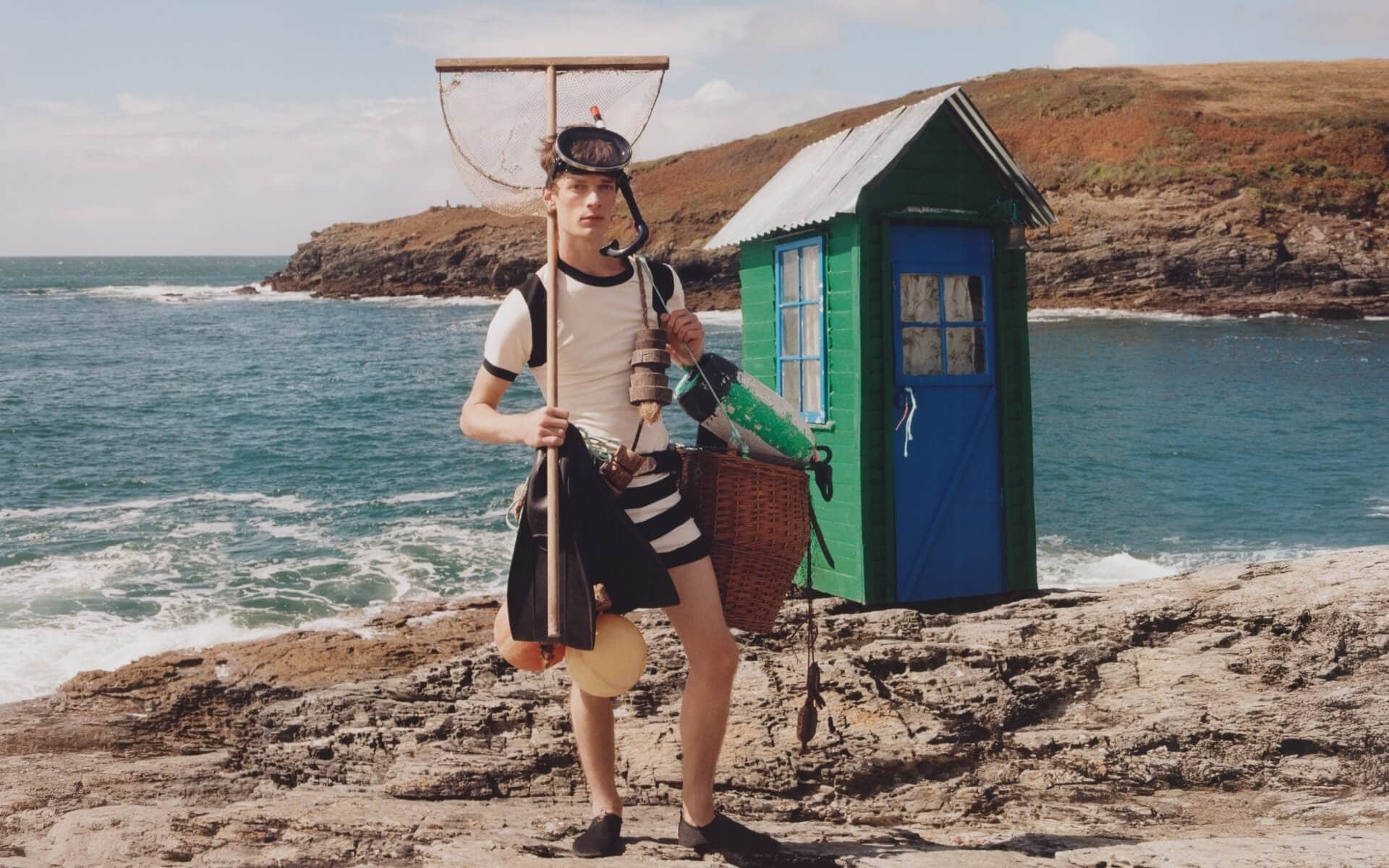 Person in striped swimwear holds fishing gear beside green hut on rocky seaside.